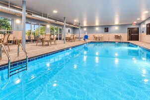 An indoor swimming pool with clear blue water, surrounded by lounge chairs and tables, and large windows offering views of greenery outside.