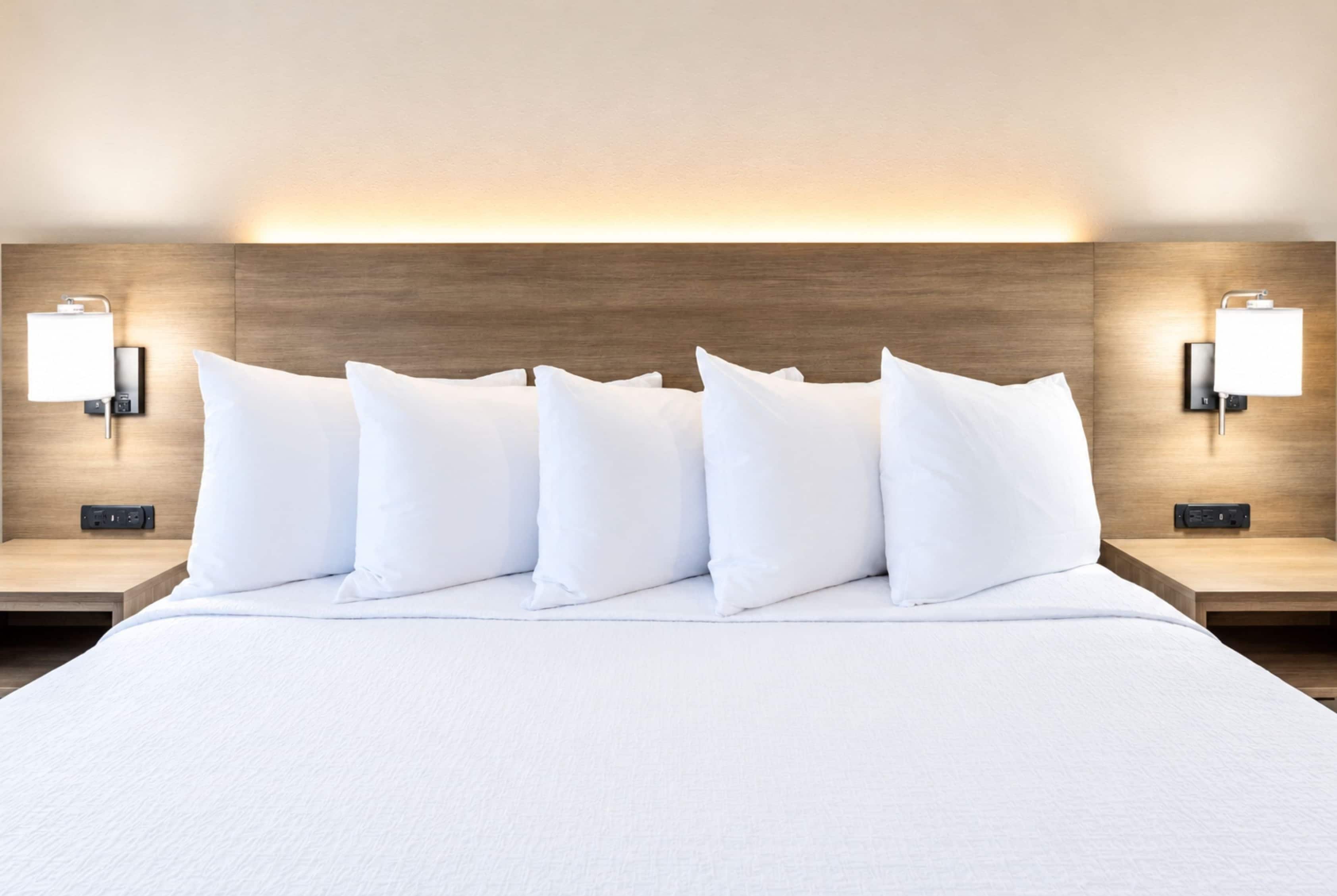 Habitación de hotel con una cama grande, varios almohadones, dos mesitas de noche y lámparas de pared.