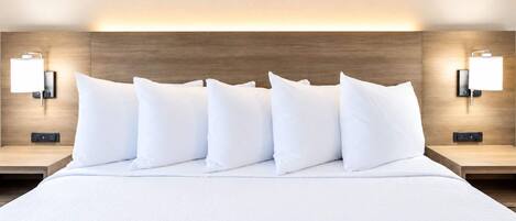 Habitación de hotel con una cama grande, varios almohadones, dos mesitas de noche y lámparas de pared.