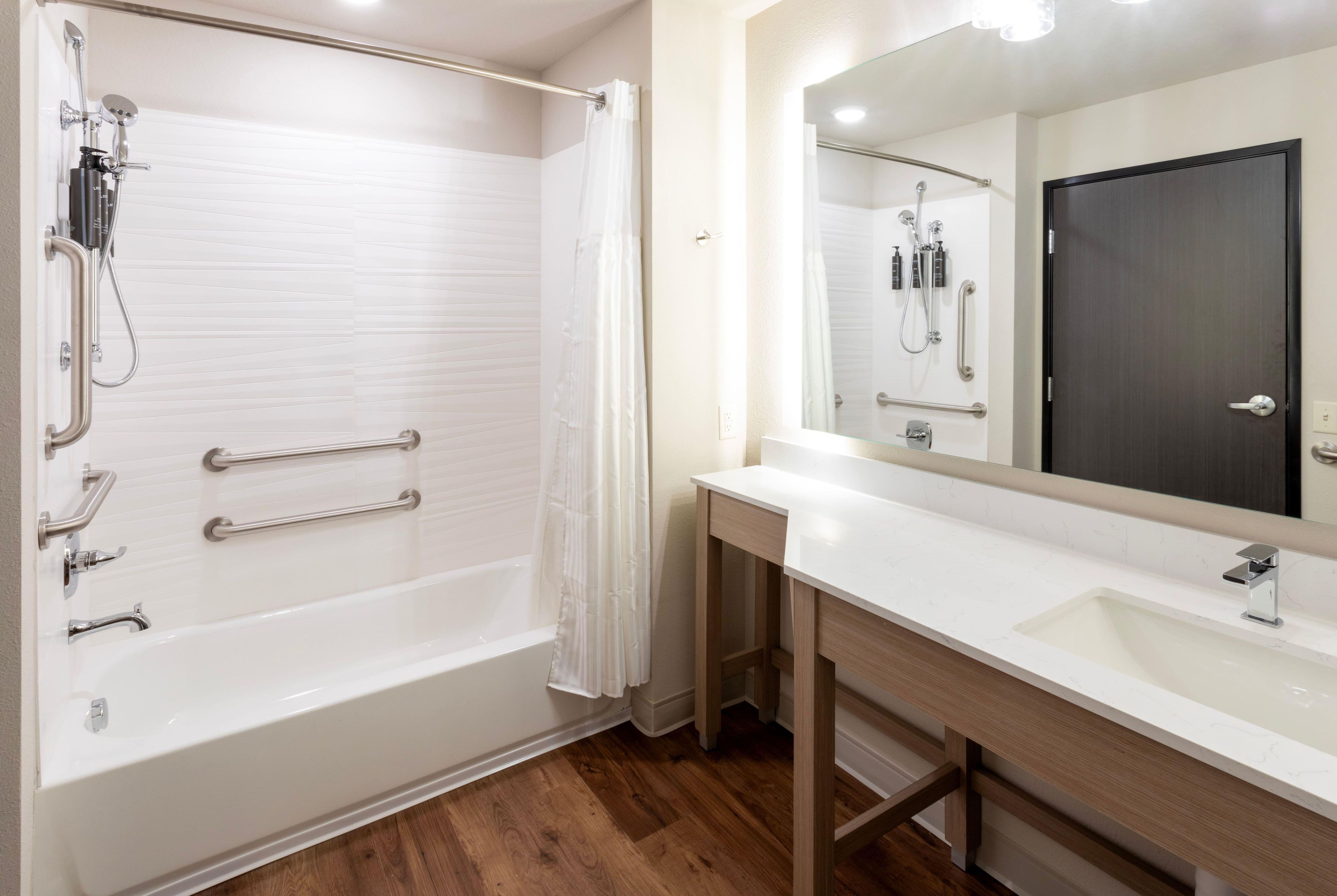 Un baño moderno con cabina de ducha, barras de apoyo, tocador con espejo y suelo de madera.