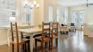 A spacious living room with a large wooden dining table, multiple chairs, and a sofa. There are large windows with blinds, a ceiling fan, and a chandelier. - Emerald Gem by Avantstay Pool Access, 3mins to Beach! (Santa Rosa Beach)