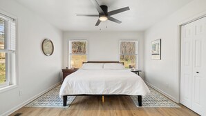 Ein Schlafzimmer mit einem Bett, einem Deckenventilator, zwei Fenstern, einer Tür und einem gemusterten Teppich.