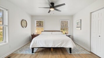 Un dormitorio con una cama, ventilador de techo, dos ventanas, una puerta y un tapete estampado.