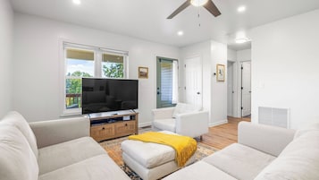 Una sala moderna con televisor de pantalla plana, sofás blancos, mueble de entretenimiento de madera y ventilador de techo.