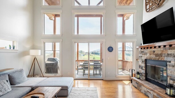 A spacious living room with a large fireplace, a sofa, a coffee table, and a television. There are multiple windows and a sliding glass door leading to an outdoor patio. - Private Retreat w/ Hot Tub, Amazing Views, Game Room Silver Spoon by Avantstay (Leicester)