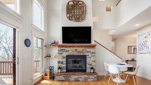 A modern living room with a stone fireplace, a flat-screen TV, and a dining area with white chairs. - Private Retreat w/ Hot Tub, Amazing Views, Game Room Silver Spoon by Avantstay (Leicester)