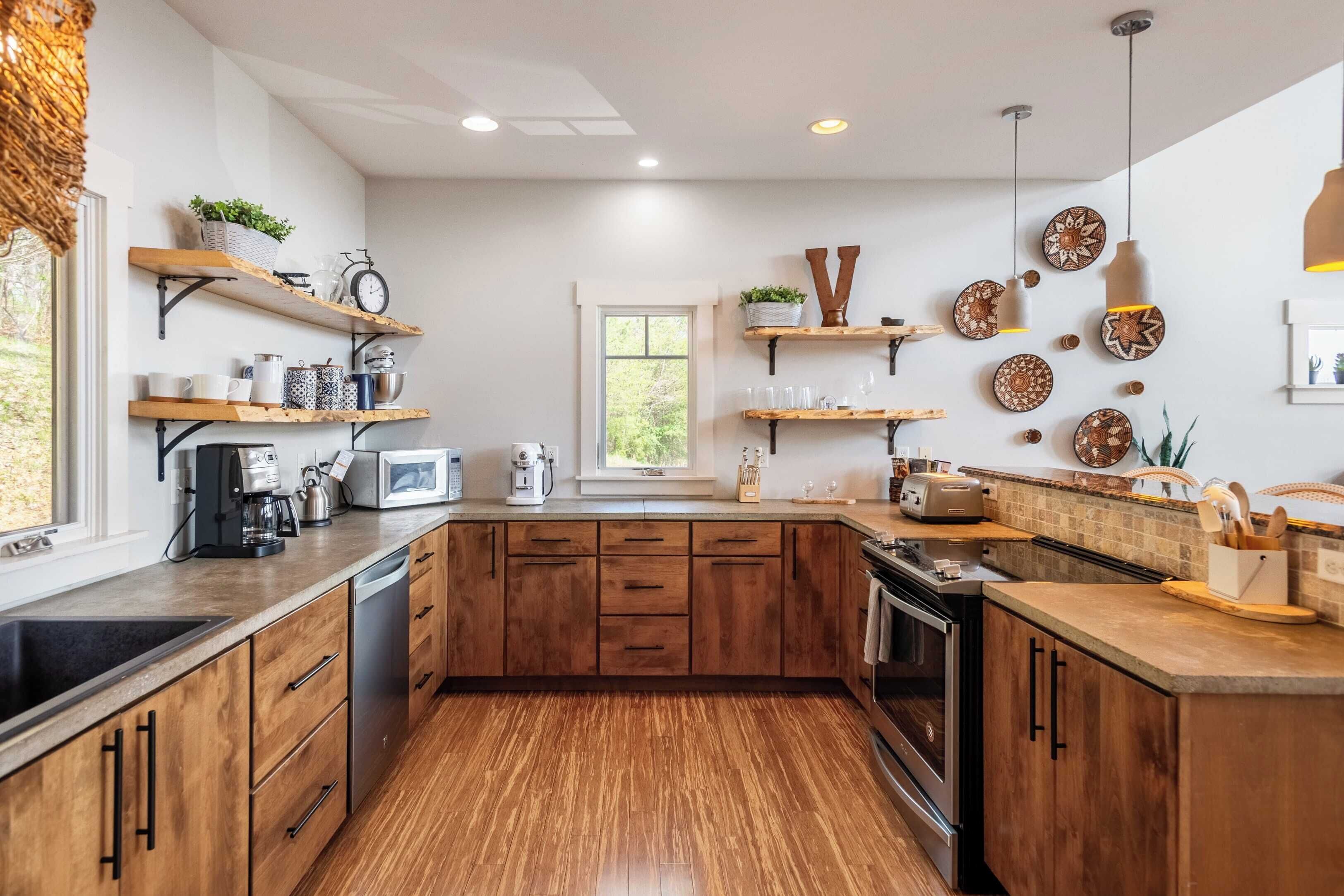 Une cuisine moderne avec des meubles en bois, un four encastré et un four à micro-ondes. On y trouve des étagères ornées d’objets décoratifs et une fenêtre qui laisse entrer la lumière naturelle.