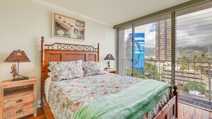 Un dormitorio con una cama grande, cabecera de madera, colcha estampada con motivos florales, dos mesitas de noche con lámparas y un ventanal con vistas a la ciudad.