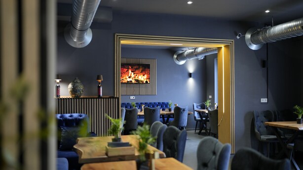 Een modern restaurant met een centraal geplaatste open haard, blauwe gestoffeerde zitplaatsen en een houten eettafel.