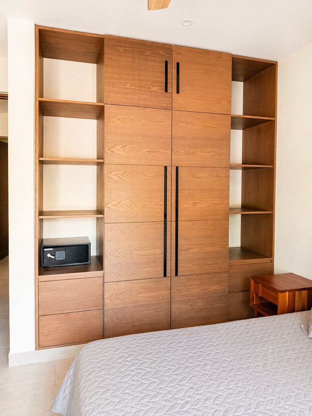 A bedroom with a bed, a wooden wardrobe with multiple shelves, a small wooden table, and a black electronic device on the wardrobe.
