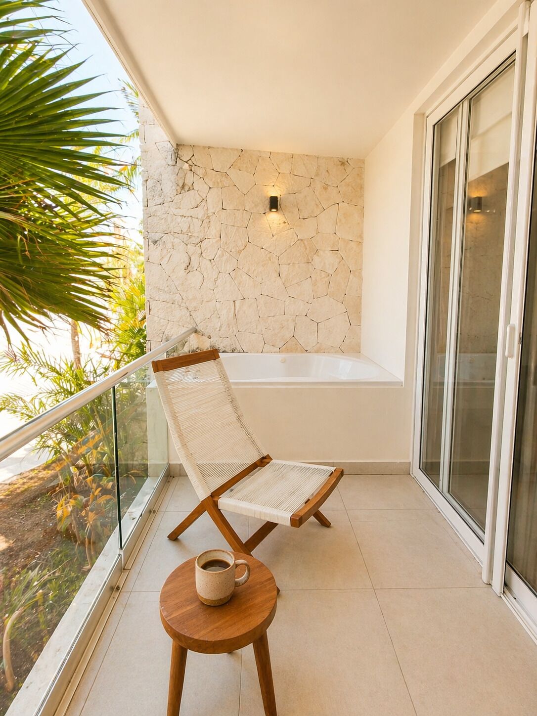 A balcony with a stone wall, a glass door, a chair, a small table, and a view of palm trees.