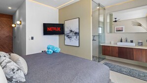A modern bedroom with a large bed, a TV mounted on the wall, a bathroom with a glass shower, and a ceiling fan.