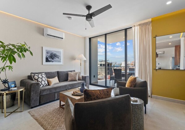 A modern living room with a grey sofa, a coffee table, and a large window offering a city view.