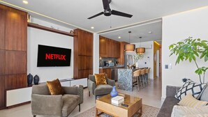 A modern living room with a flat-screen TV, sofas, a coffee table, and a kitchen in the background.
