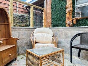 A wicker chair with a cushioned backrest and a glass-top table, positioned in front of a stone wall with a metal gate and a wooden post. - 3 bedroom accommodation in Ashover, near Matlock. (Ashover, near Matlock.)