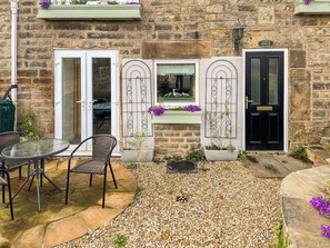 A stone building with a black door, a white door, and a window with decorative ironwork. There is a table and two chairs outside. - 3 bedroom accommodation in Ashover, near Matlock. (Ashover, near Matlock.)