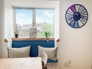 A room with a wooden table, a blue bench with cushions, a window with a view of buildings, and a wall clock. - 3 bedroom accommodation in Ashover, near Matlock. (Ashover, near Matlock.)
