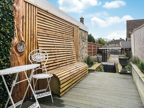 A wooden deck with a bench and two white metal tables, surrounded by greenery and a stone wall.