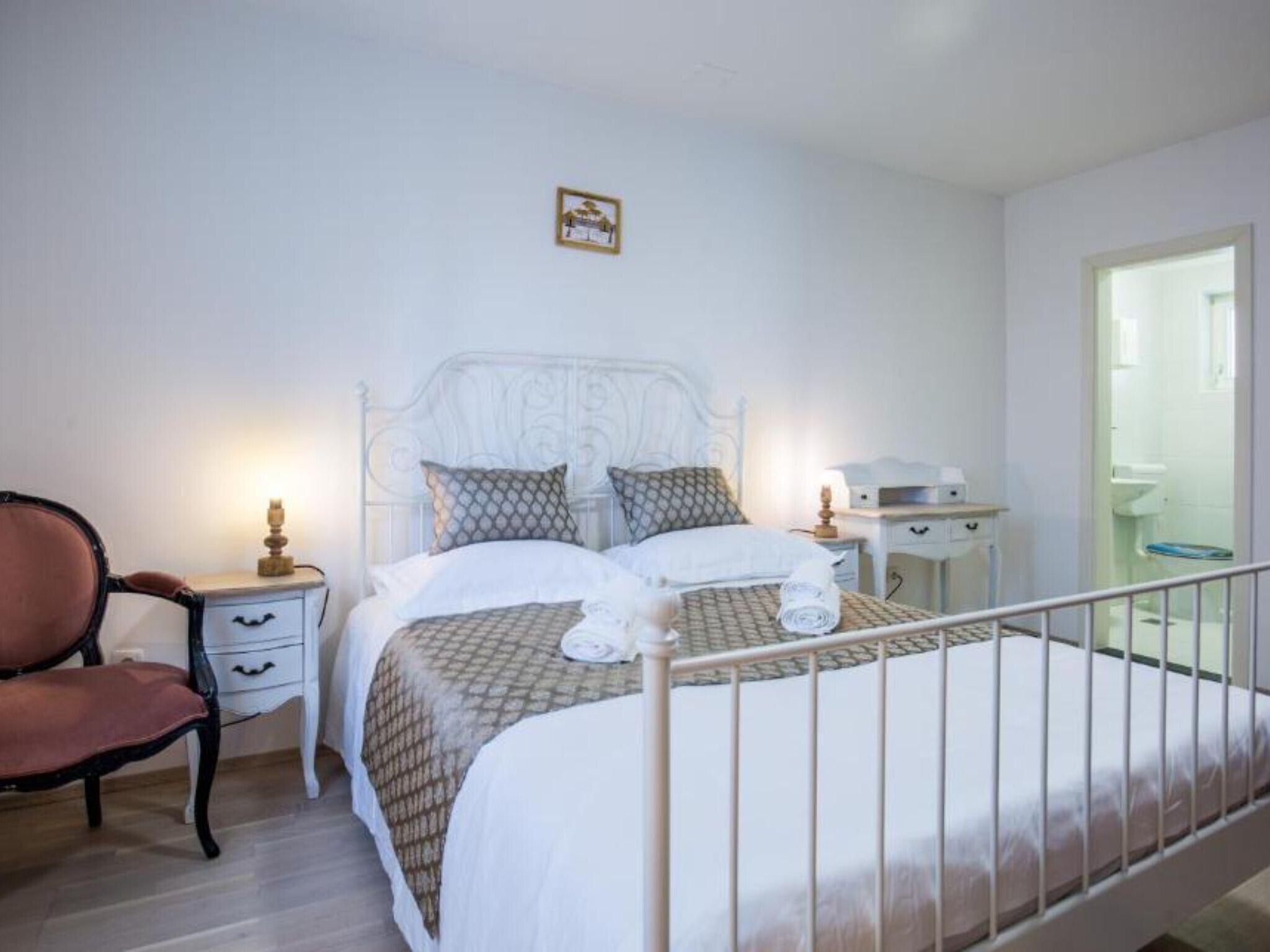 A neatly arranged bedroom with a bed, bedside tables, a chair, and a door leading to a bathroom.