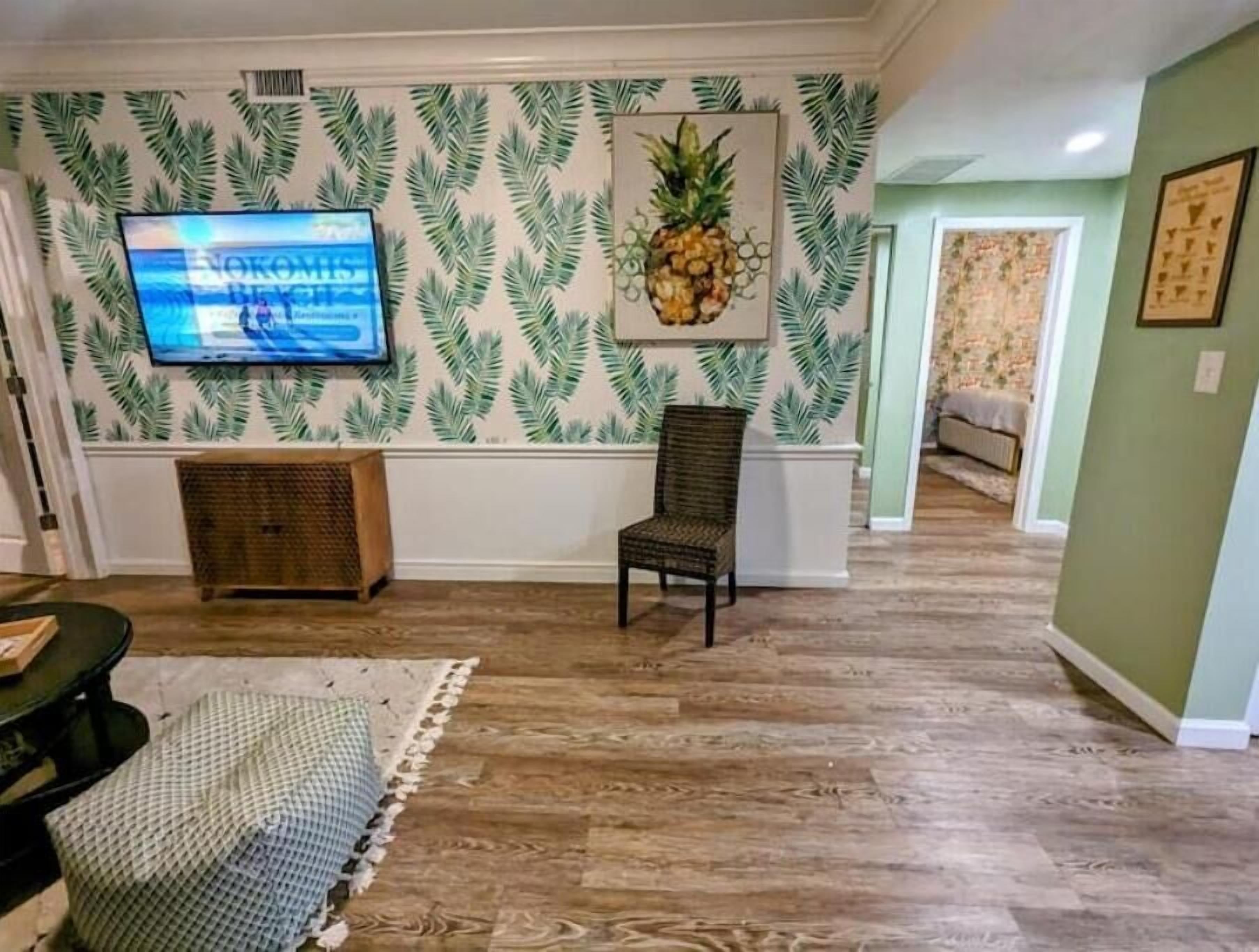 A living room with a television, a wicker bench, a patterned rug, and a pineapple artwork on the wall.