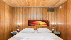 A wooden-paneled room with a bed, a painting of a sunset, and a bench with a colorful blanket.