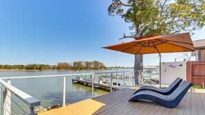 Een houten terras met loungestoelen en een parasol, met uitzicht op een kalm wateroppervlak en aangemeerde boten.