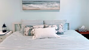 Een slaapkamer met een bed, voorzien van gestreept beddengoed, een kussen met een kaartmotief en een schilderij van een uitzicht op de oceaan erboven.
