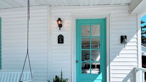 Een wit huis met een turquoise deur, een schommel op het terras en een kleine bloempot.