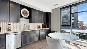 A modern kitchen with dark cabinets, a dishwasher, and a round glass table with a white chair. - Rare Luxury: Apartment with Unparalleled Elegance (Rochester)