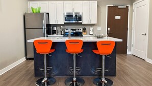 A modern kitchen with white cabinets, a stainless steel refrigerator, a microwave, and a gas stove. There are three orange bar stools and a small island with a sink. - Bright, Colorful & Convenient 1-Bedroom Apartment (Rochester)