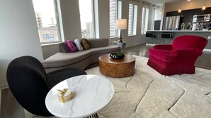 A modern living room with a sofa, a red armchair, a round coffee table, and a dining area with a kitchen in the background. - Tower280 LUXE One-Bedroom Corner Unit with Views! (Rochester)