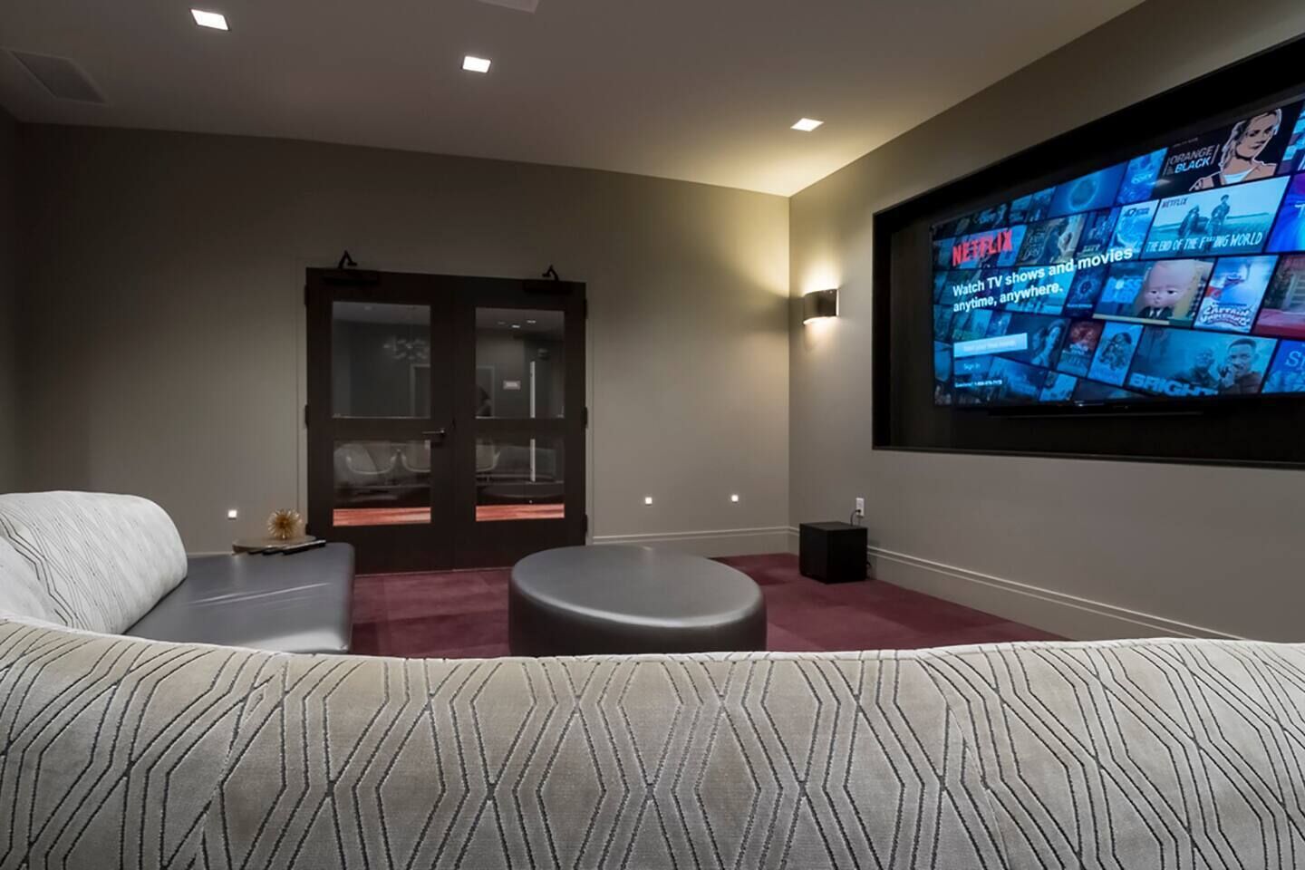 Une salle de cinéma maison moderne, dotée d’un grand écran diffusant du contenu Netflix, d’un espace salon confortable avec un canapé et un pouf, et d’une porte donnant sur une autre pièce.