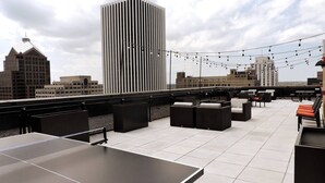 Une terrasse sur le toit avec des sièges en plein air, une table de ping-pong et des guirlandes lumineuses, le tout avec la silhouette de la ville en toile de fond.