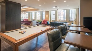 Une table de billard avec des chaises et une table de jeu de flipper dans une pièce dotée de grandes fenêtres et offrant une vue sur les bâtiments environnants.