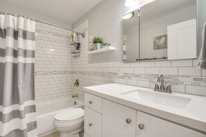 A bathroom with a white sink, a toilet, a bathtub, and a shower curtain. There are shelves above the sink with toiletries. - Charming Orem Home w/ Yard - Near BYU & UVU! (Orem)