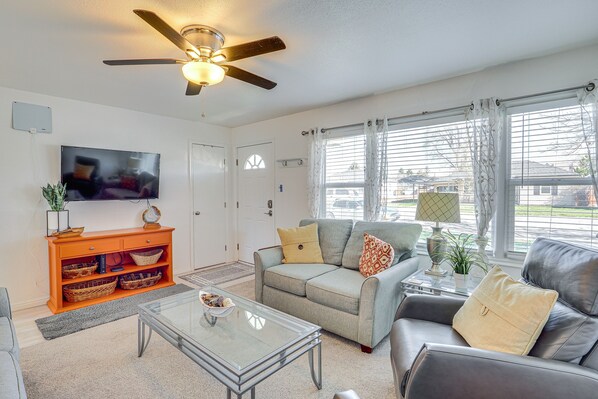 A living room with a ceiling fan, a flat-screen TV, a sofa, a coffee table, and a window with curtains. - Charming Orem Home w/ Yard - Near BYU & UVU! (Orem)