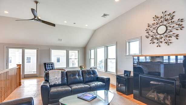 A spacious living room with a leather sofa, a glass coffee table, a fireplace, and large windows. - Narragansett Home w/ Scenic Views: Walk to Beach! (Narragansett)