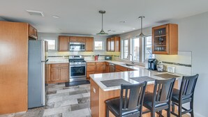 A modern kitchen with wooden cabinets, a central island, and a dining area with chairs. - Narragansett Home w/ Scenic Views: Walk to Beach! (Narragansett)