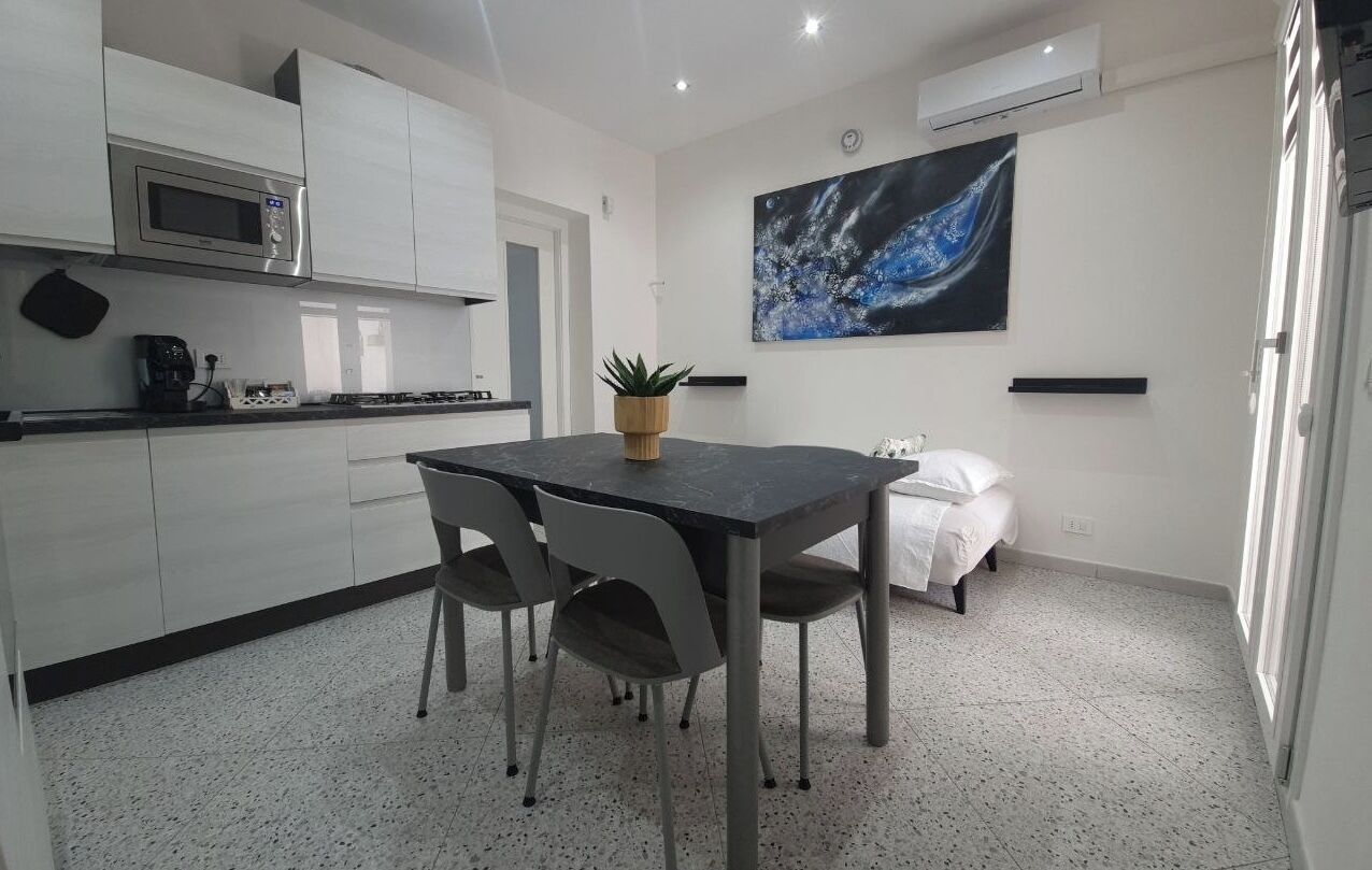 Una cocina moderna con armarios blancos, un microondas y una cafetera. Hay una mesa de comedor con sillas y una planta en maceta. Un cuadro adorna la pared, y un sofá blanco se encuentra en la esquina.
