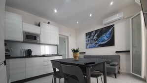 A modern kitchen with white cabinets, a microwave, and a stove. There is a dining table with chairs and a grey sofa. A large abstract painting is on the wall, and an air conditioning unit is mounted on the wall. - Private House 14 Palermo (Palermo)