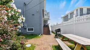 Un jardin avec un chemin en briques, une table de pique-nique en bois, un barbecue et une clôture blanche.