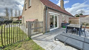 Une maison en briques avec un toit recouvert de tuiles rouges, une clôture en bois et une terrasse avec une table et des chaises.