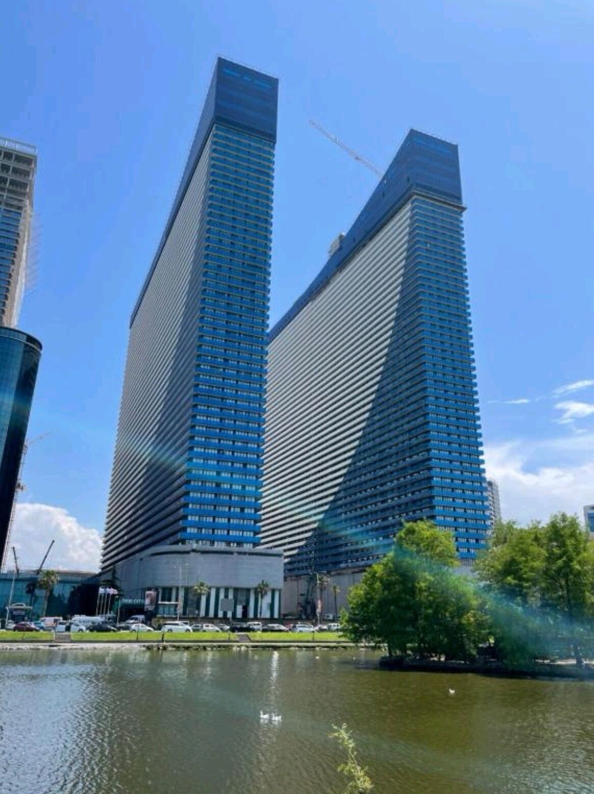 Two tall, modern skyscrapers with reflective glass facades, a body of water in the foreground, and a small island with trees.