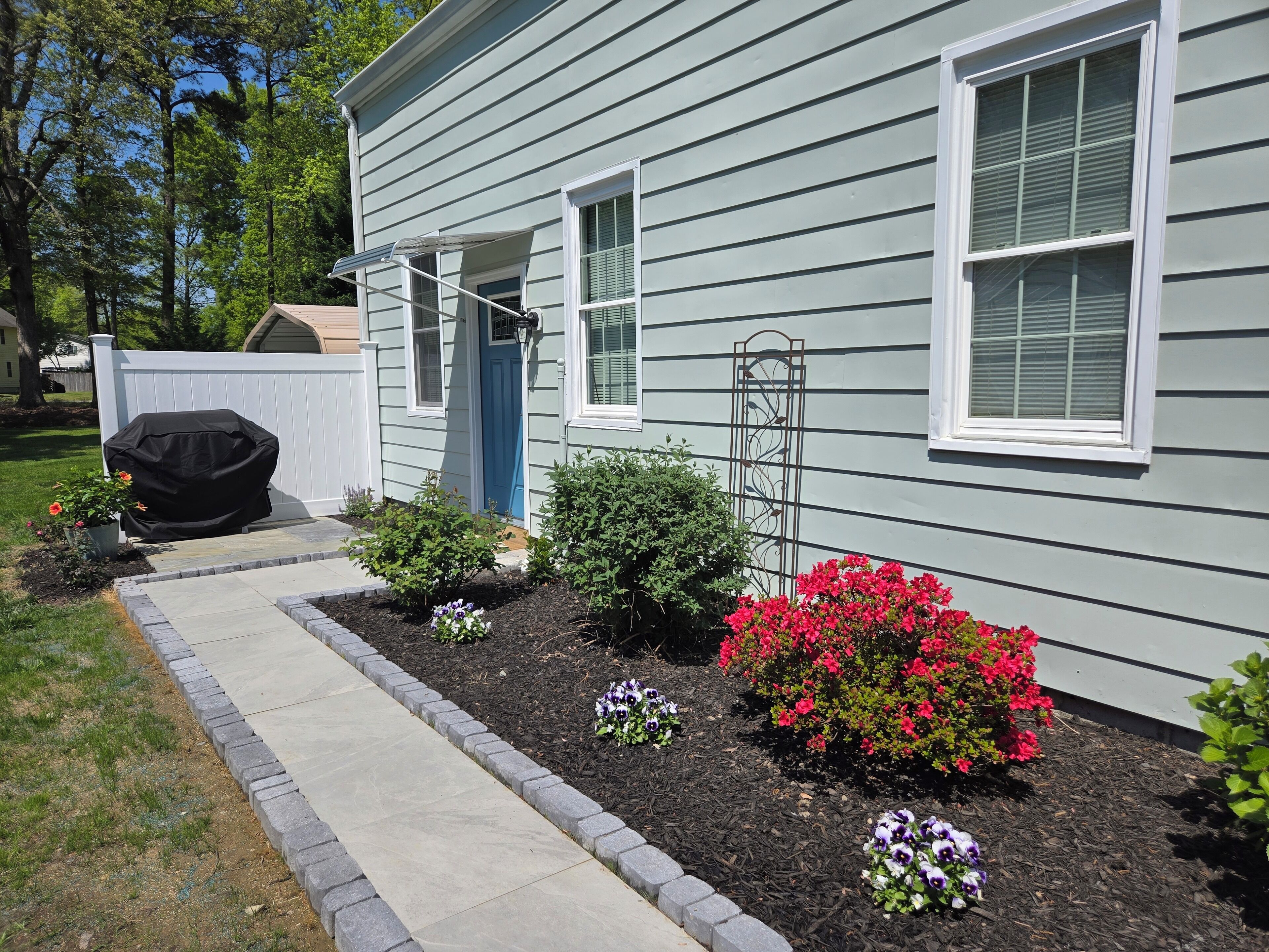 A house with a covered grill, a pathway, and flower beds with various flowers.