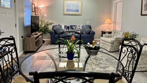 Ein Wohnzimmer mit Glastisch, blauem Sofa, gemustertem Sessel und Fernseher auf einem Holzboden.