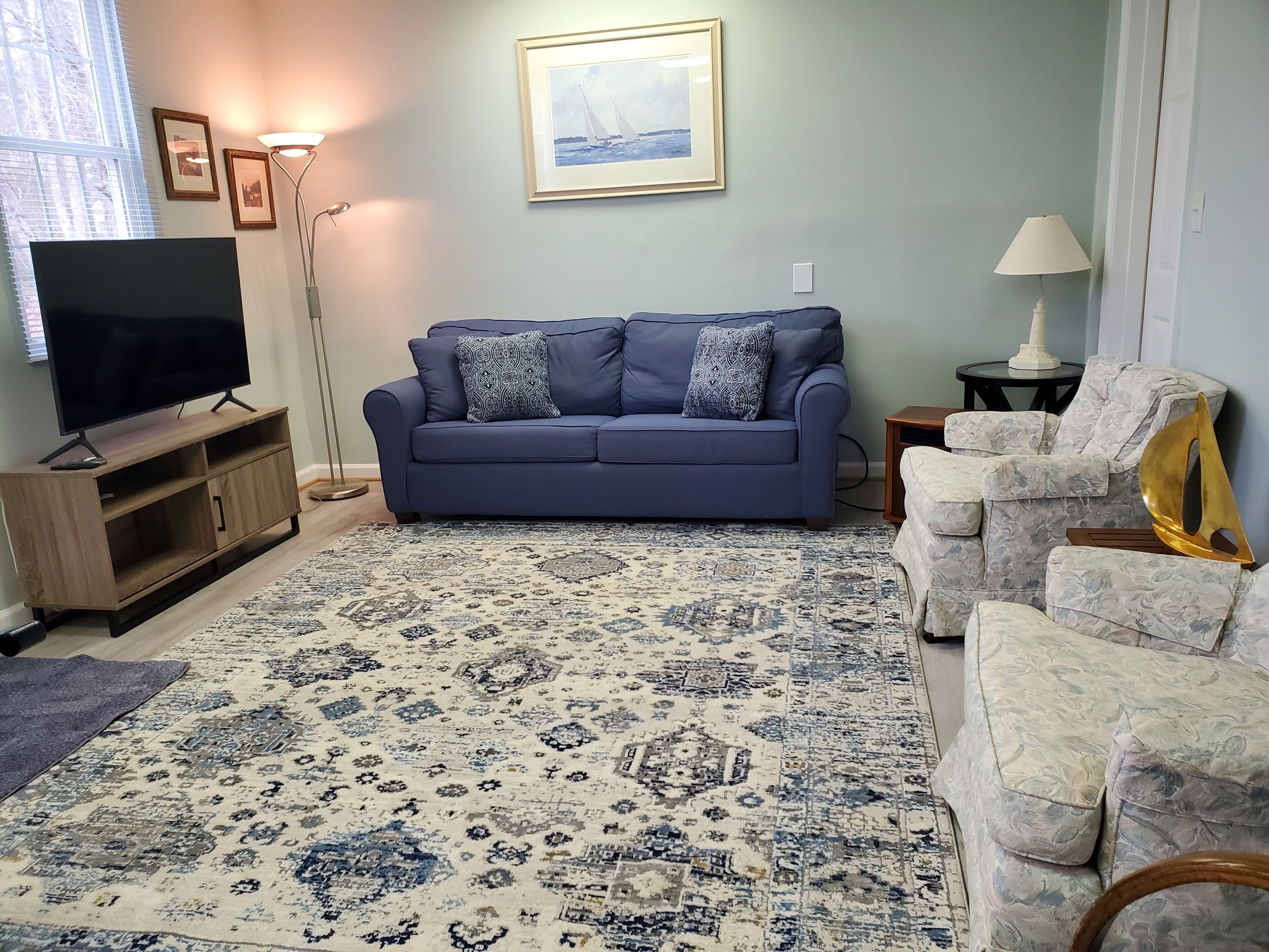 Un salon avec un canapé bleu, un téléviseur posé sur un meuble en bois, un tapis à motifs et un fauteuil à fleurs.
