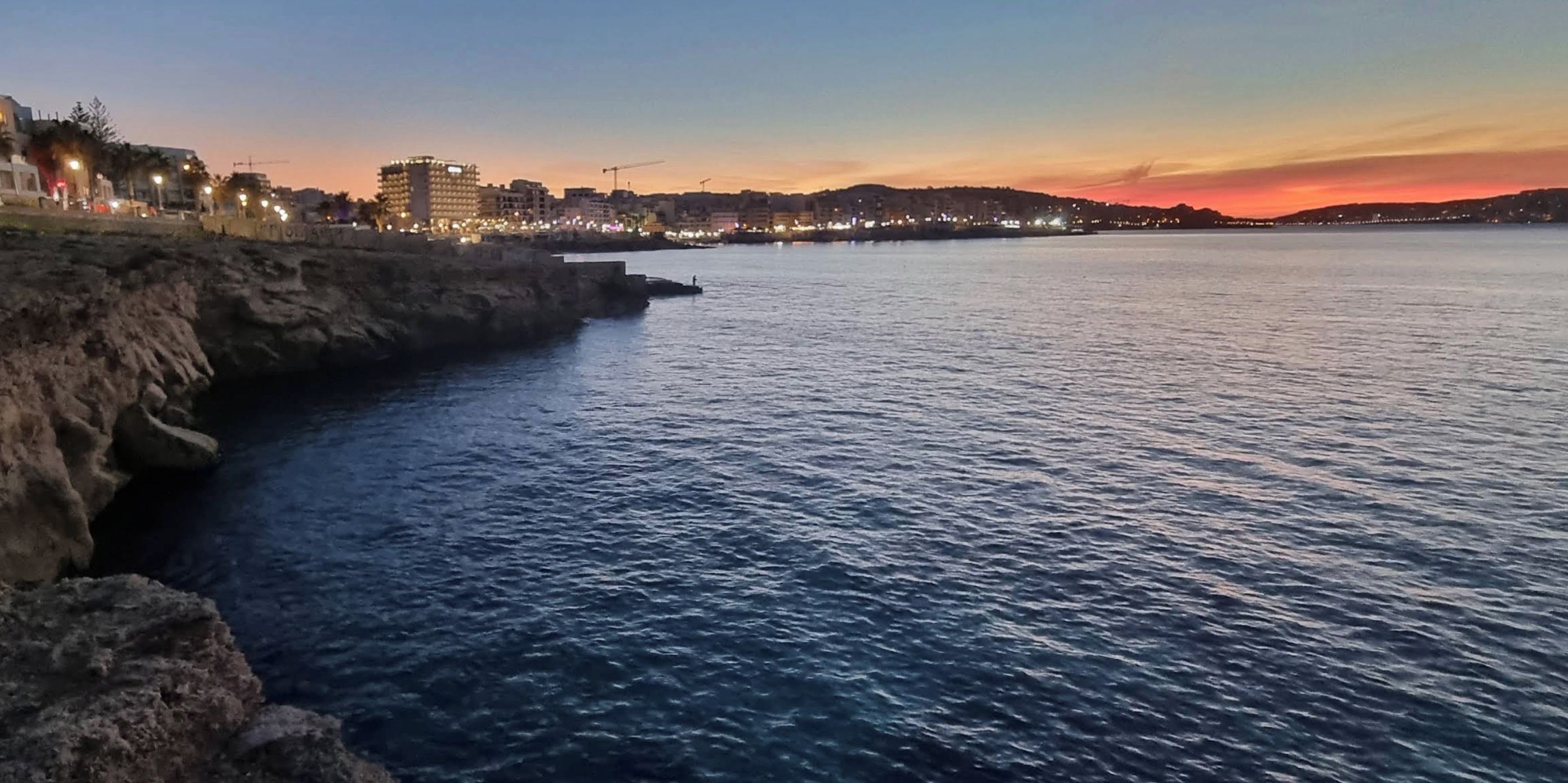 A coastal cityscape at dusk with buildings along the shore and a calm sea.
