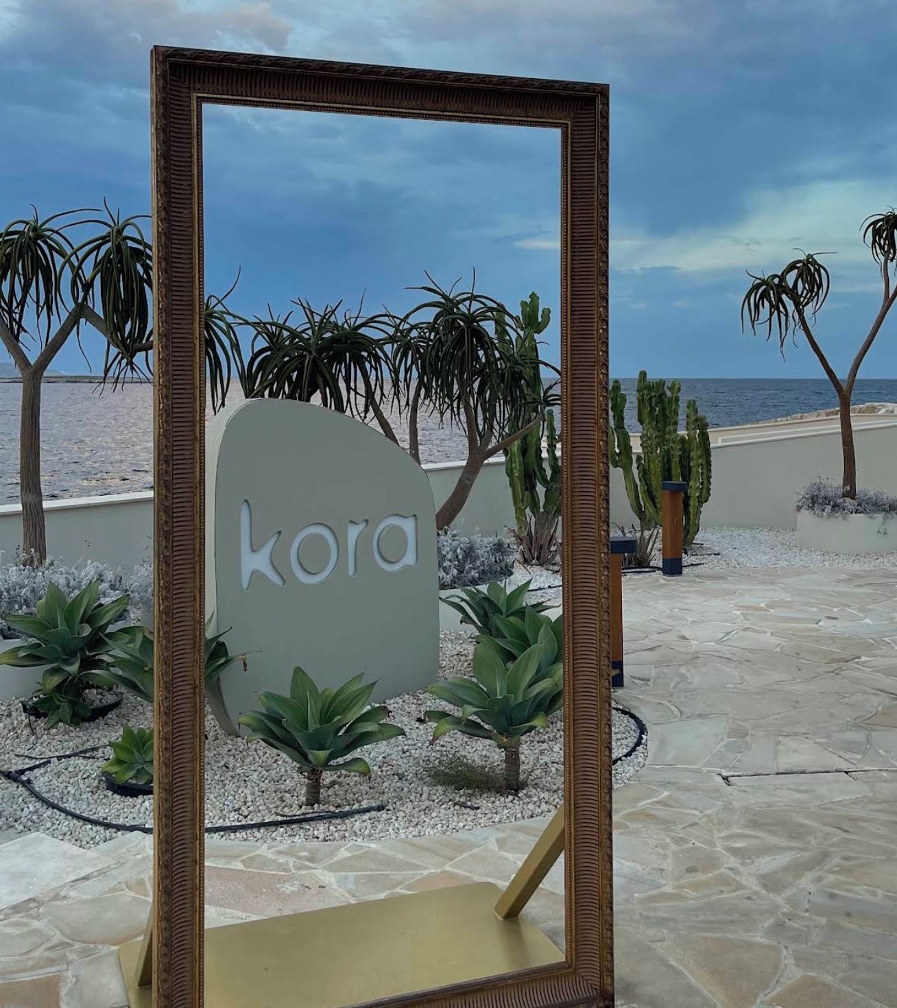 Ein gerahmtes Schild mit dem Wort 'kora' in einer wüstenähnlichen Landschaft mit Kakteen und Palmen.