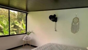 Une chambre avec une grande fenêtre, un lit recouvert d’une couvre-lit à motifs géométriques, une petite table ronde avec une plante et deux éléments de décoration murale.