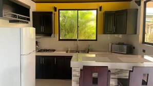 Une cuisine avec des meubles de couleur sombre, un réfrigérateur blanc, une fenêtre donnant sur un espace verdoyant et un sol carrelé.